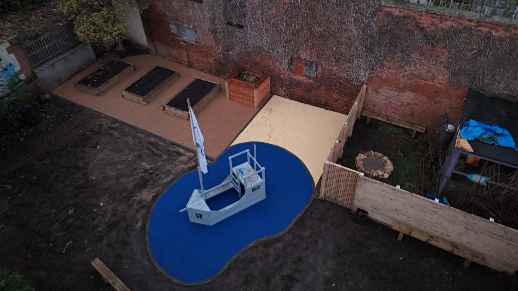 Aerial view of the finished community play garden, with a wooden play boat on safety surfacing, a sand pit and oak raised beds in a walled garden