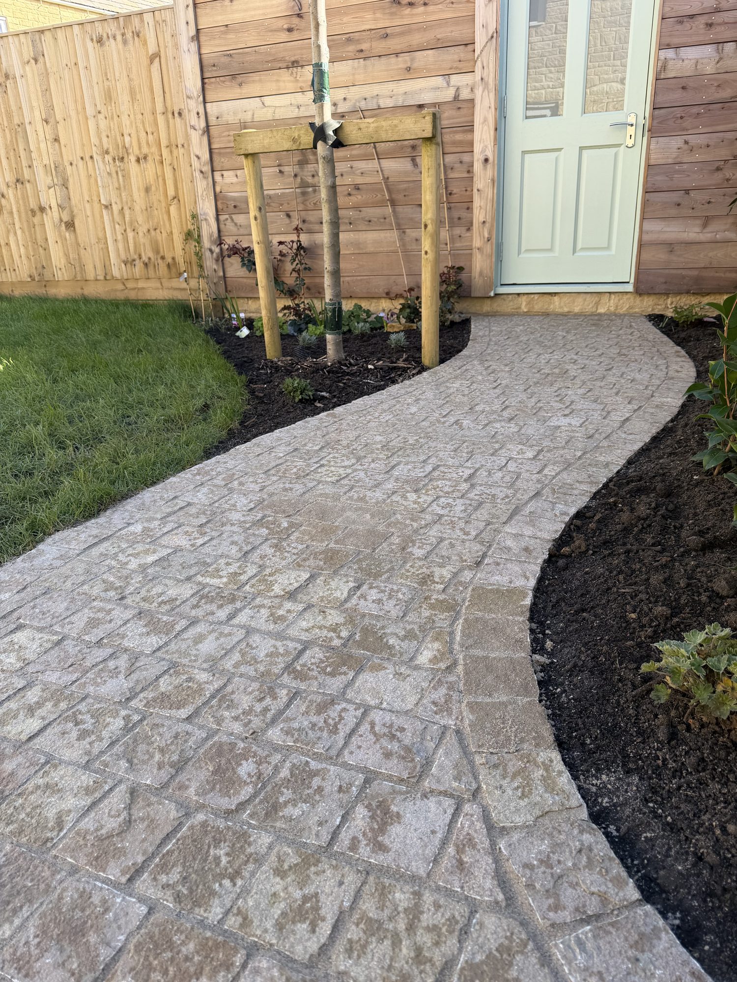 Tumbled sett path winding to a sage-green cottage door with new planting and a young tree